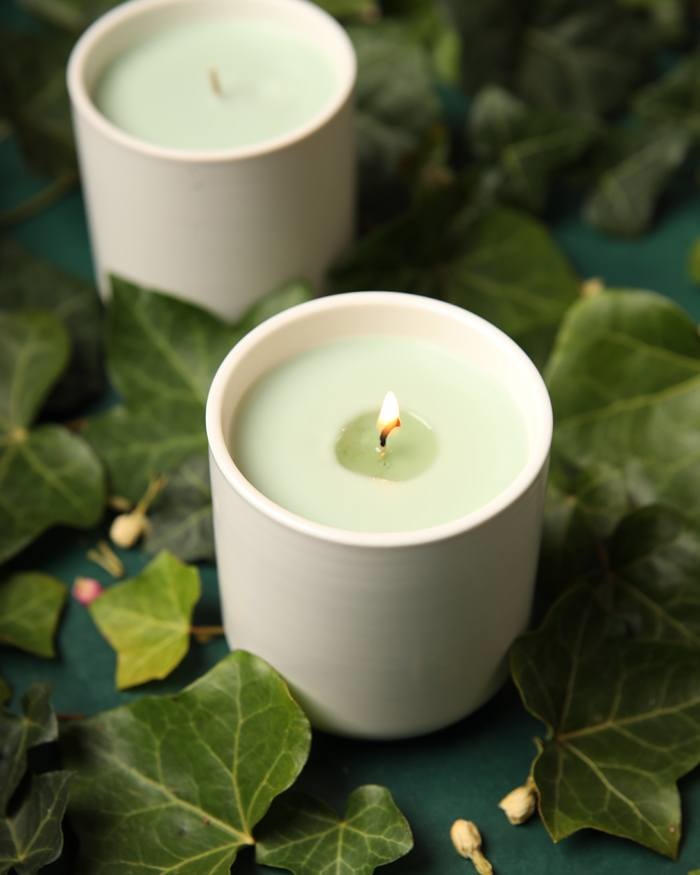A green candle in a white jar burning on a table among ivy leaves